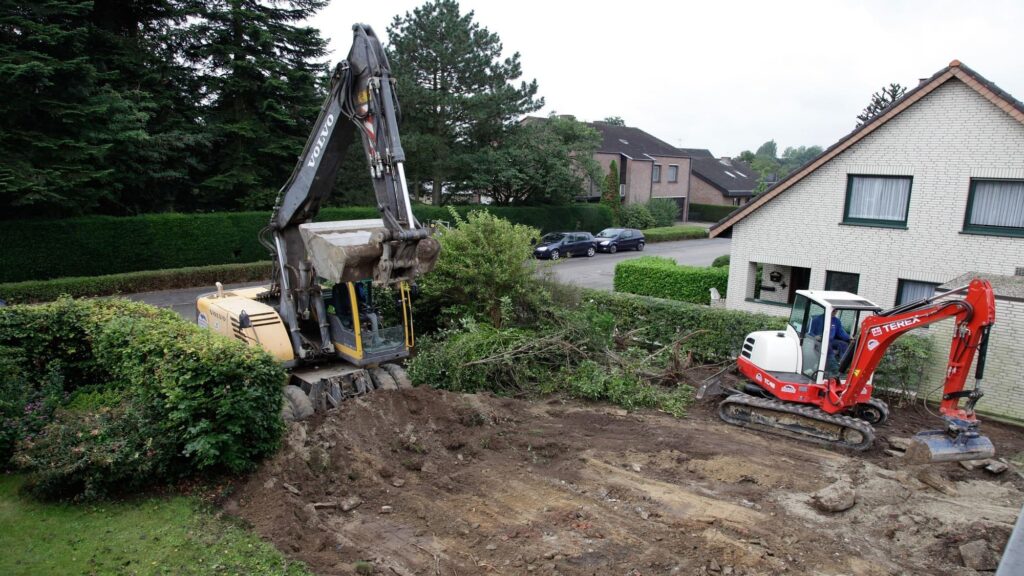 Vue aérienne de deux pelleteuses en action devant une maison résidentielle : à gauche, une pelleteuse Volvo jaune sur pneus ; à droite, une mini-pelleteuse Terex rouge sur chenilles, en train de creuser et dégager le sol bordé de haies et de véhicules garés.