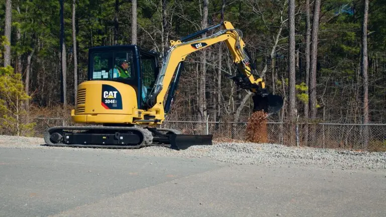 Mini-pelle CAT 304E2 en action sur un chantier de terrassement en bord de route &agrave; Gen&egrave;ve, avec op&eacute;rateur en s&eacute;curit&eacute;, en train de d&eacute;placer des mat&eacute;riaux gravillonn&eacute;s sur terrain stabilis&eacute;.